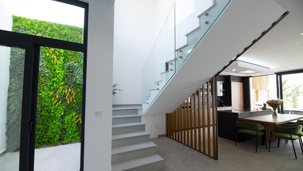 Un interior moderno con una escalera que conduce a un segundo piso, un comedor con una mesa y sillas de madera, y vista a un jardín verde a través de un vidrio.