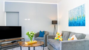 Ett modernt vardagsrum med en platt-TV, ett soffbord med en vas med blommor, en grå soffa med färgglada kuddar och en blå, abstrakt målning på väggen.