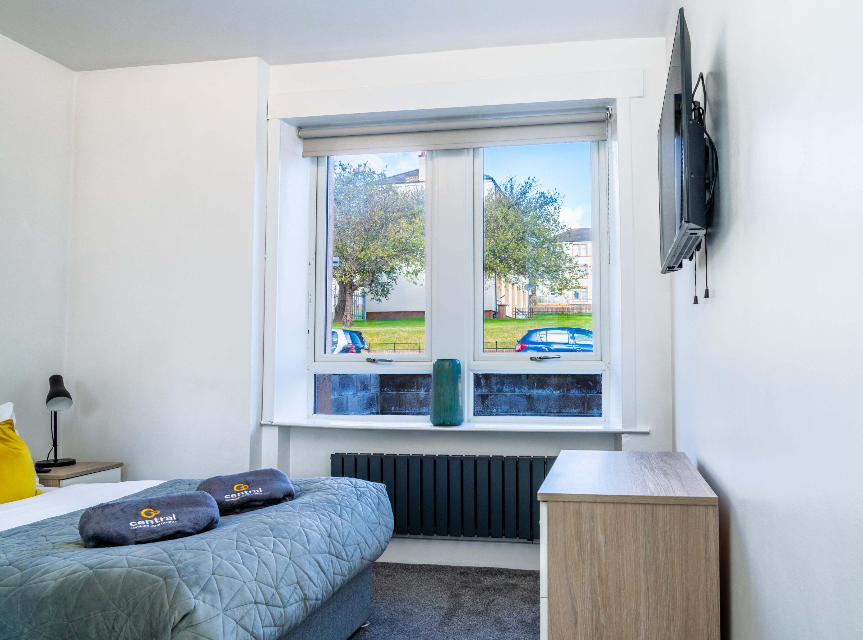 Een slaapkamer met een bed, een nachtkastje, een lamp, een radiator, een raam met uitzicht op de straat en een aan de muur bevestigde televisie.