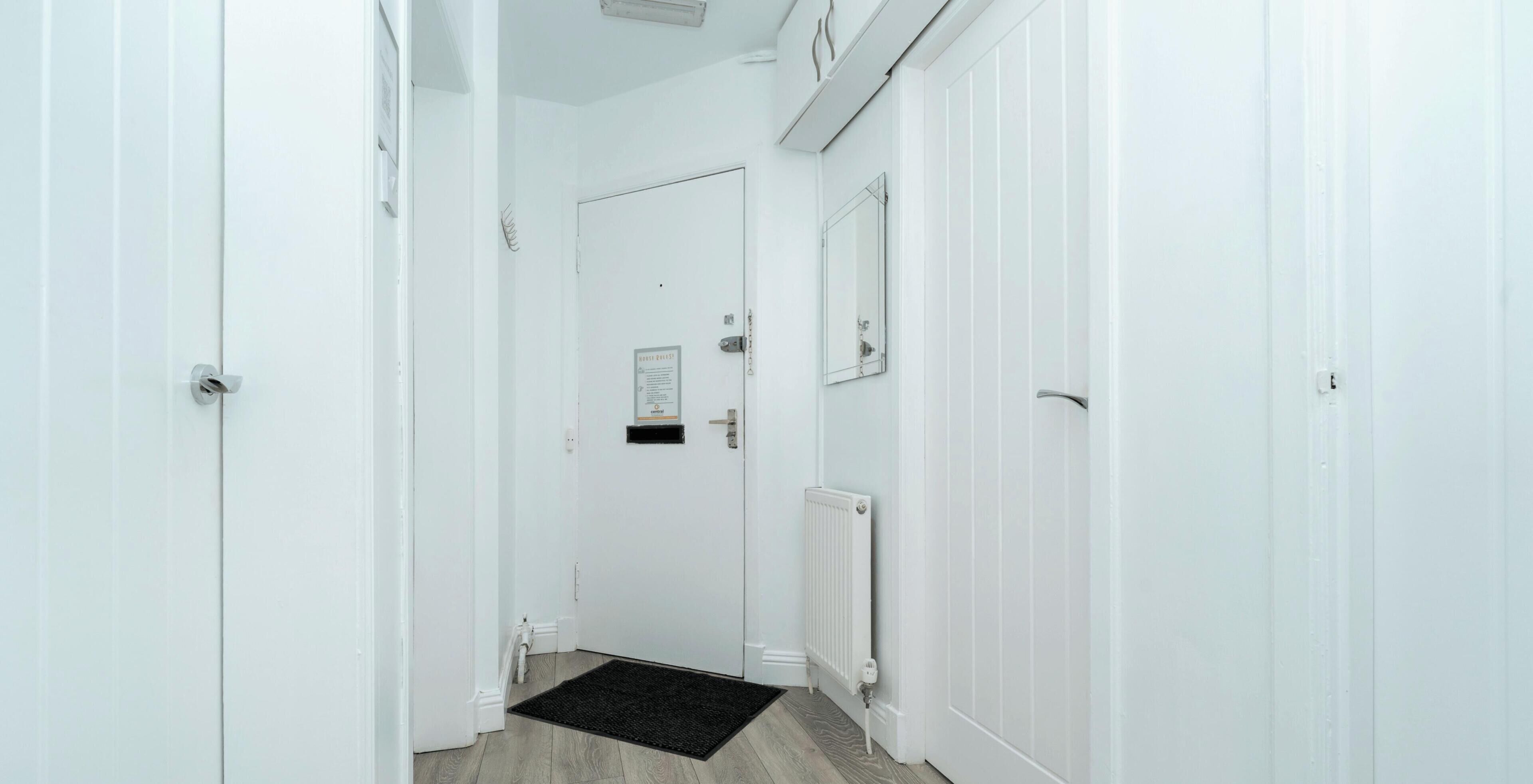 A narrow hallway with white walls, a black doormat, a door with a sign, and a radiator.