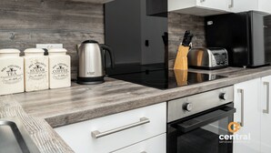 A modern kitchen with white cabinets, a wooden backsplash, a black induction cooktop, a toaster, and a coffee maker. - Trades Lane by Central Serviced Apartments - City Centre (Dundee)