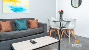 A modern living room with a grey sofa, a coffee table, and a dining area with a glass table and chairs. - Trades Lane by Central Serviced Apartments - City Centre (Dundee)