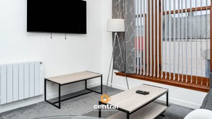 A modern living room with a flat-screen TV mounted on the wall, a floor lamp, a small table, and a window with vertical blinds. - Trades Lane by Central Serviced Apartments - City Centre (Dundee)