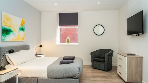 A modern bedroom with a bed, bedside tables, a chair, a TV, and a window with a view of a brick wall. - The Bond by Central Serviced Apartments - City Centre (Dundee)