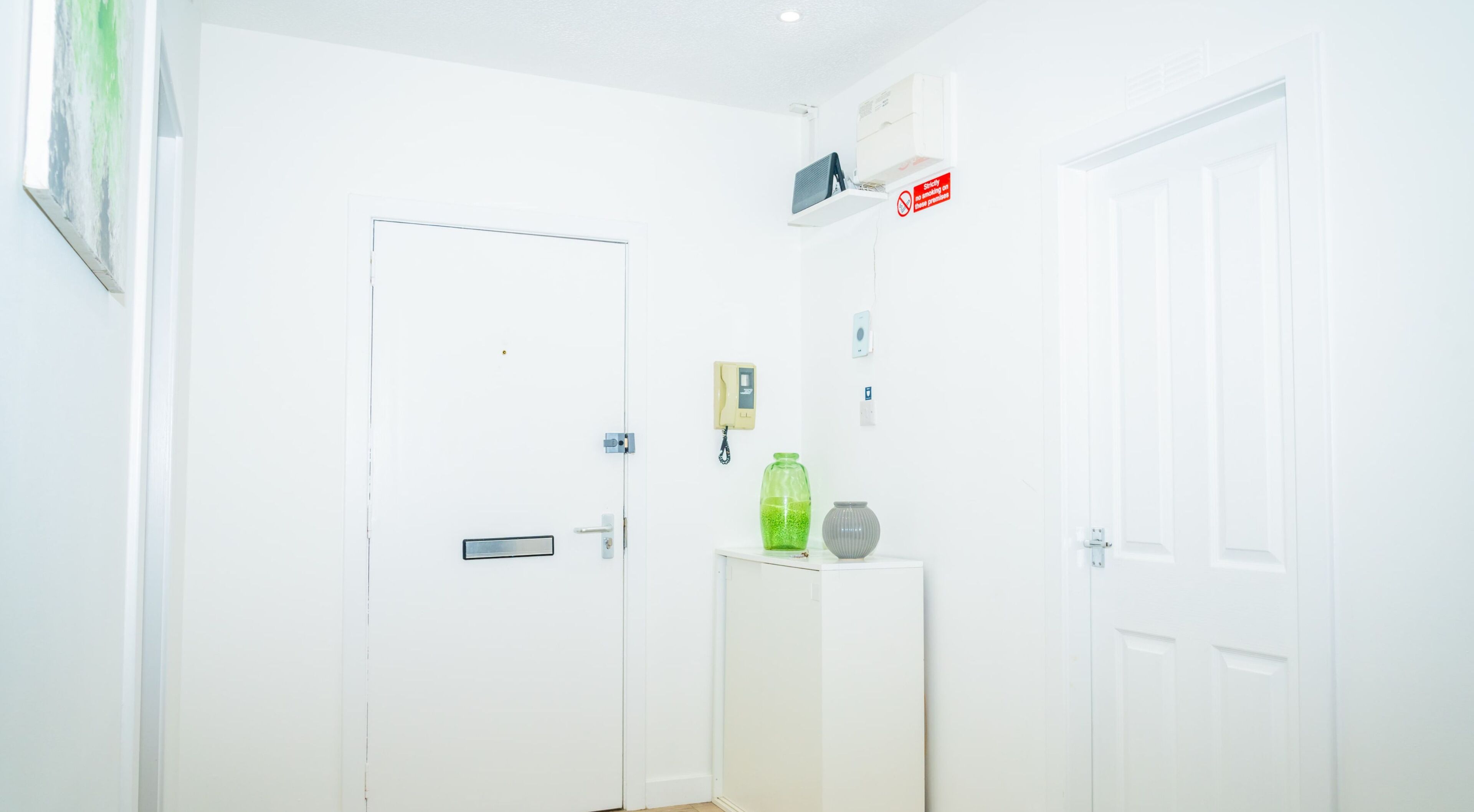 A clean, white room with a door, a small table with a green bottle, and a red no-smoking sign on the wall.