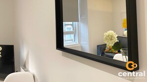 Ett modernt rum med en stor spegel, en vit stol, ett litet bord med en vas med blommor och ett fönster med utsikt över byggnader.