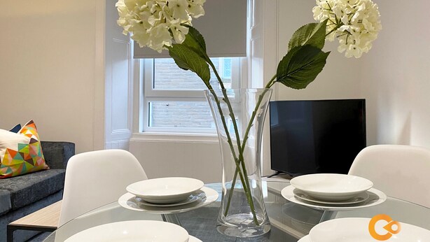 En matplats med ett bord dukat för fyra personer, två vaser med vita blommor, en tv och ett fönster med persienner.