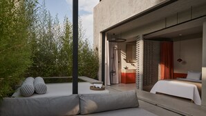 A modern outdoor seating area with a sofa and cushions, a small round table with a glass, and a view of a bedroom with a bed and a bathroom. - The Newel Psychiko (Athens)