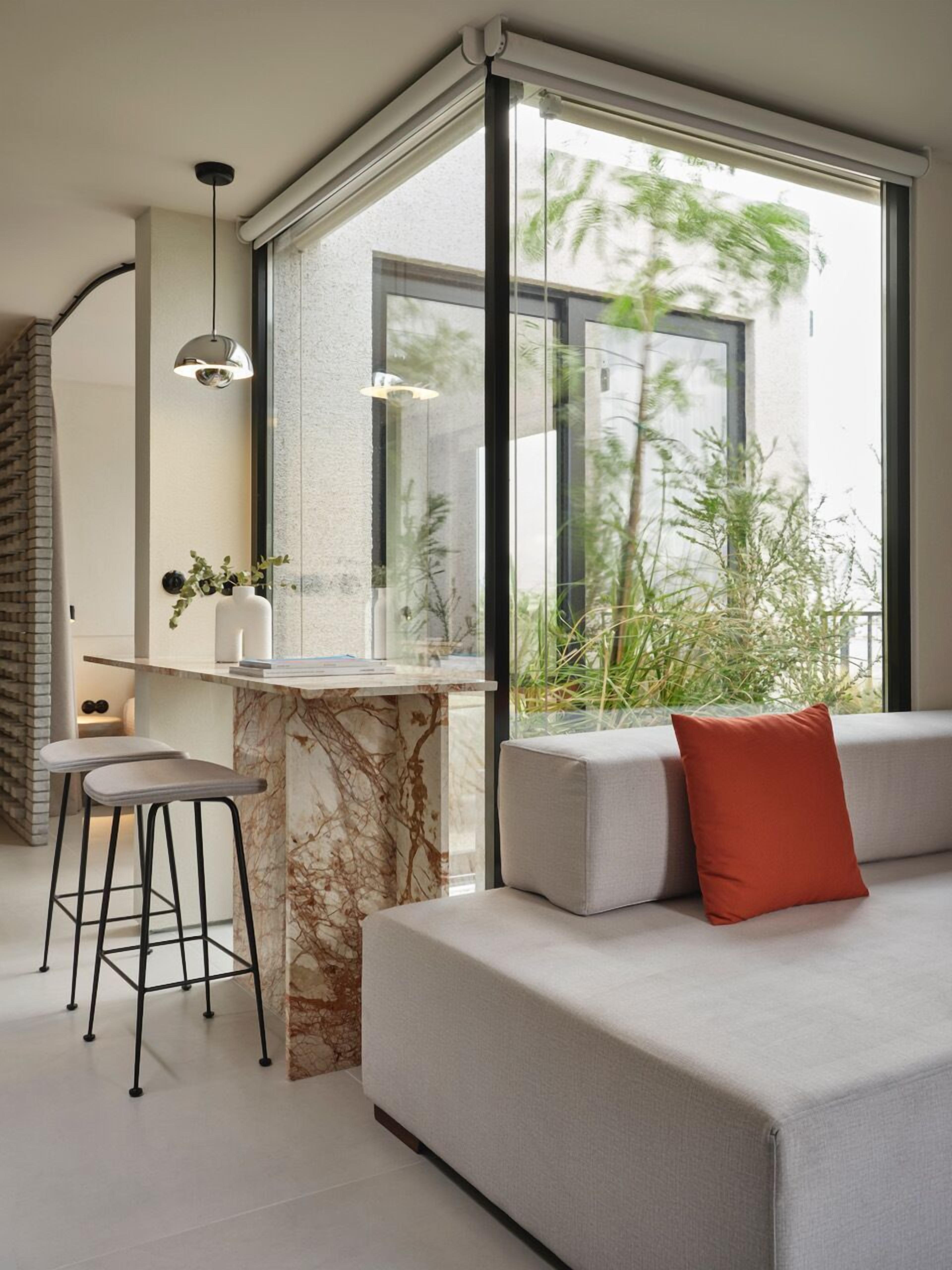 Una sala de estar moderna con una pared de acento de mármol, un sofá con un cojín rojo, un área de comedor con una mesa de mármol y ventanas grandes con vistas a zonas verdes.