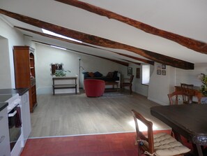 A spacious living area with a dining table, red armchair, and a sofa. There is a kitchen area with cabinets and a countertop. The room has exposed wooden beams on the ceiling and a sloped ceiling.