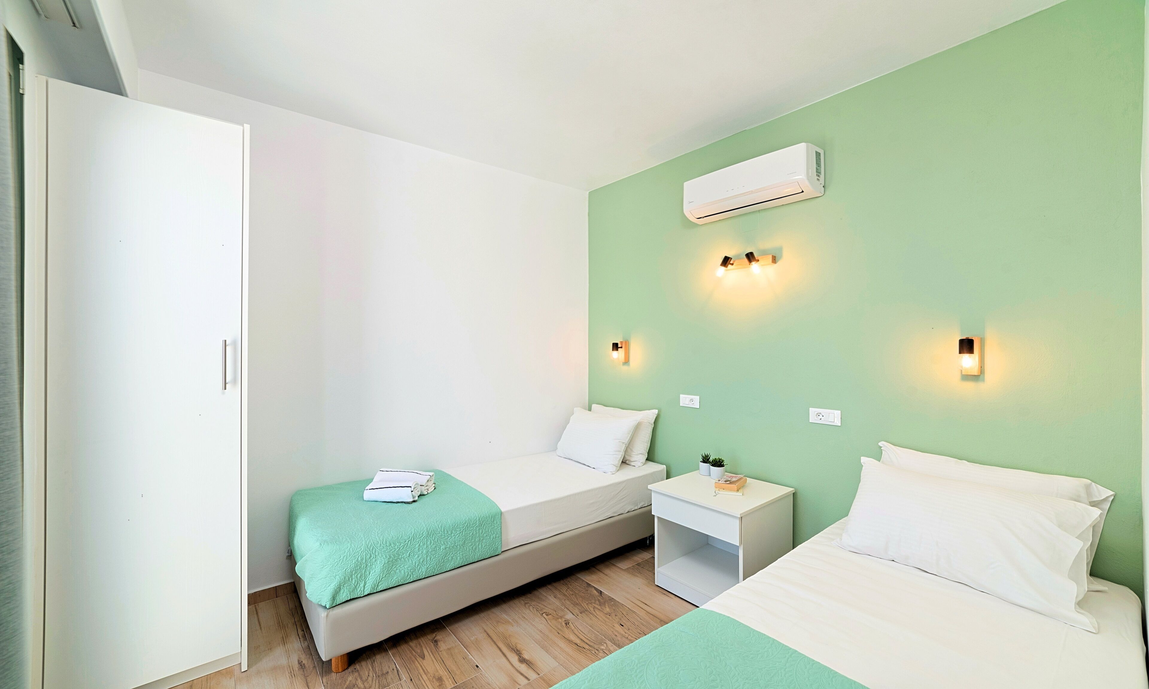 A bedroom with two beds, a wardrobe, an air conditioner, bedside lamps, and a small bedside table with a plant.