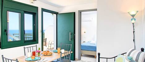 A room with a dining table set for breakfast, a bed with a blue bedspread, and a view of the sea through the windows.