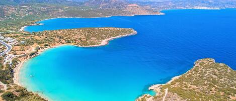 Vue aérienne d’une zone côtière avec des eaux d’un bleu limpide, une petite île et un paysage montagneux en arrière-plan.