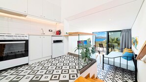A modern kitchen with white cabinets, a built-in oven, and a coffee machine. There is a balcony with a view of the sea and a dining area with a table and chairs. - Istron Homes (Agios Nikolaos)