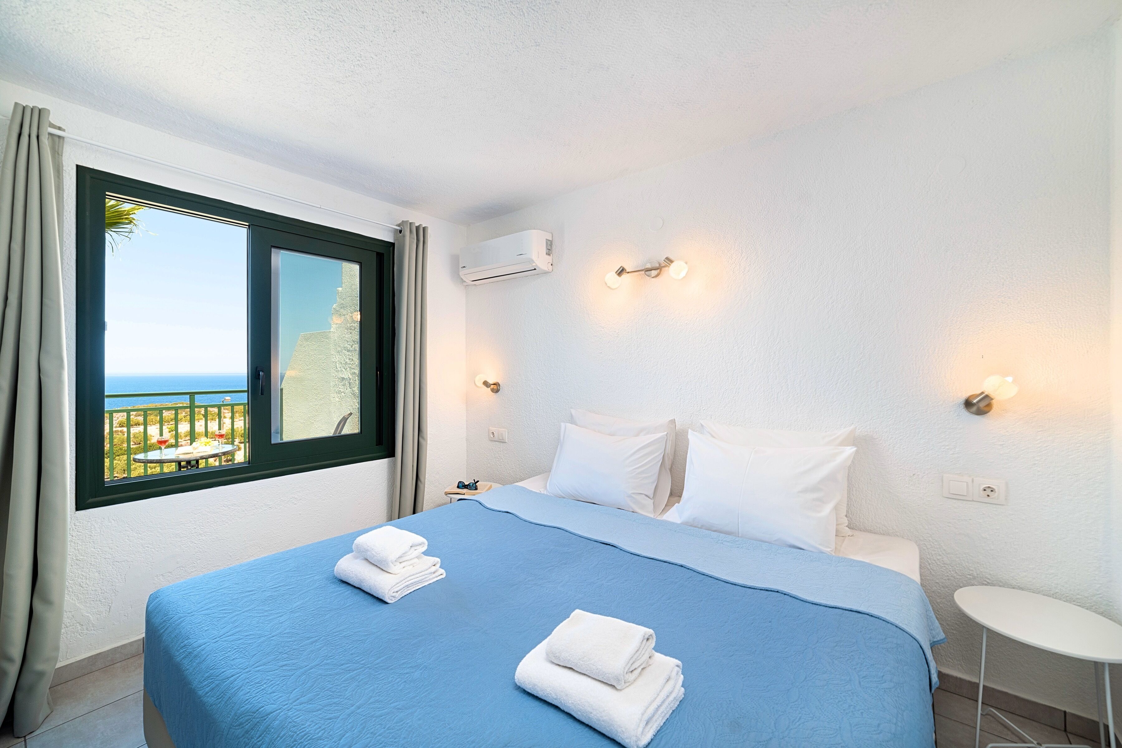 A hotel room with a bed, a small table, a window with curtains, an air conditioning unit, and a wall-mounted light fixture.