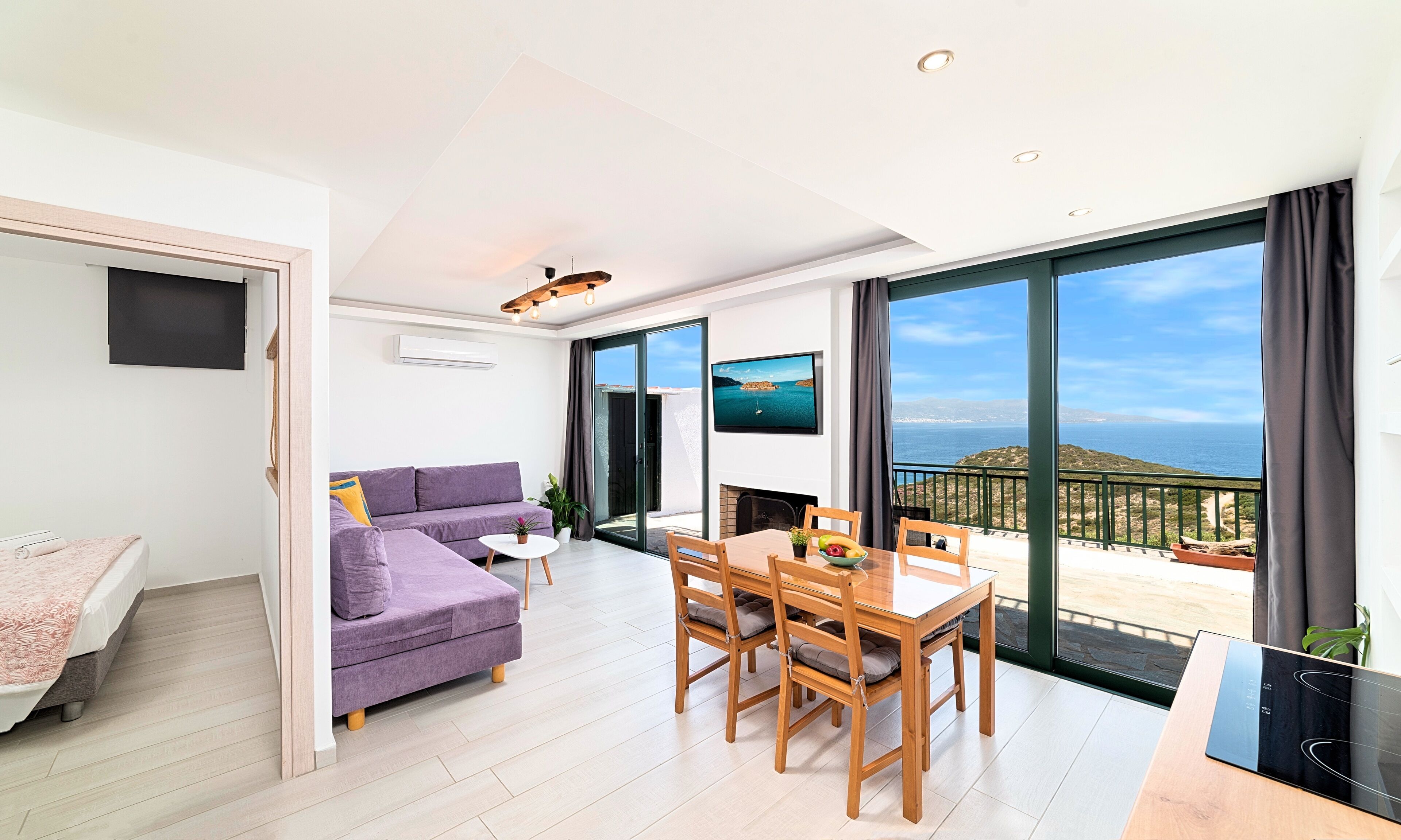 Una sala de estar moderna con sofá, mesa de comedor, sillas y televisor. Hay un balcón con vistas al mar.