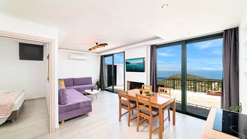 Una sala de estar moderna con sofá, mesa de comedor, sillas y televisor. Hay un balcón con vistas al mar.