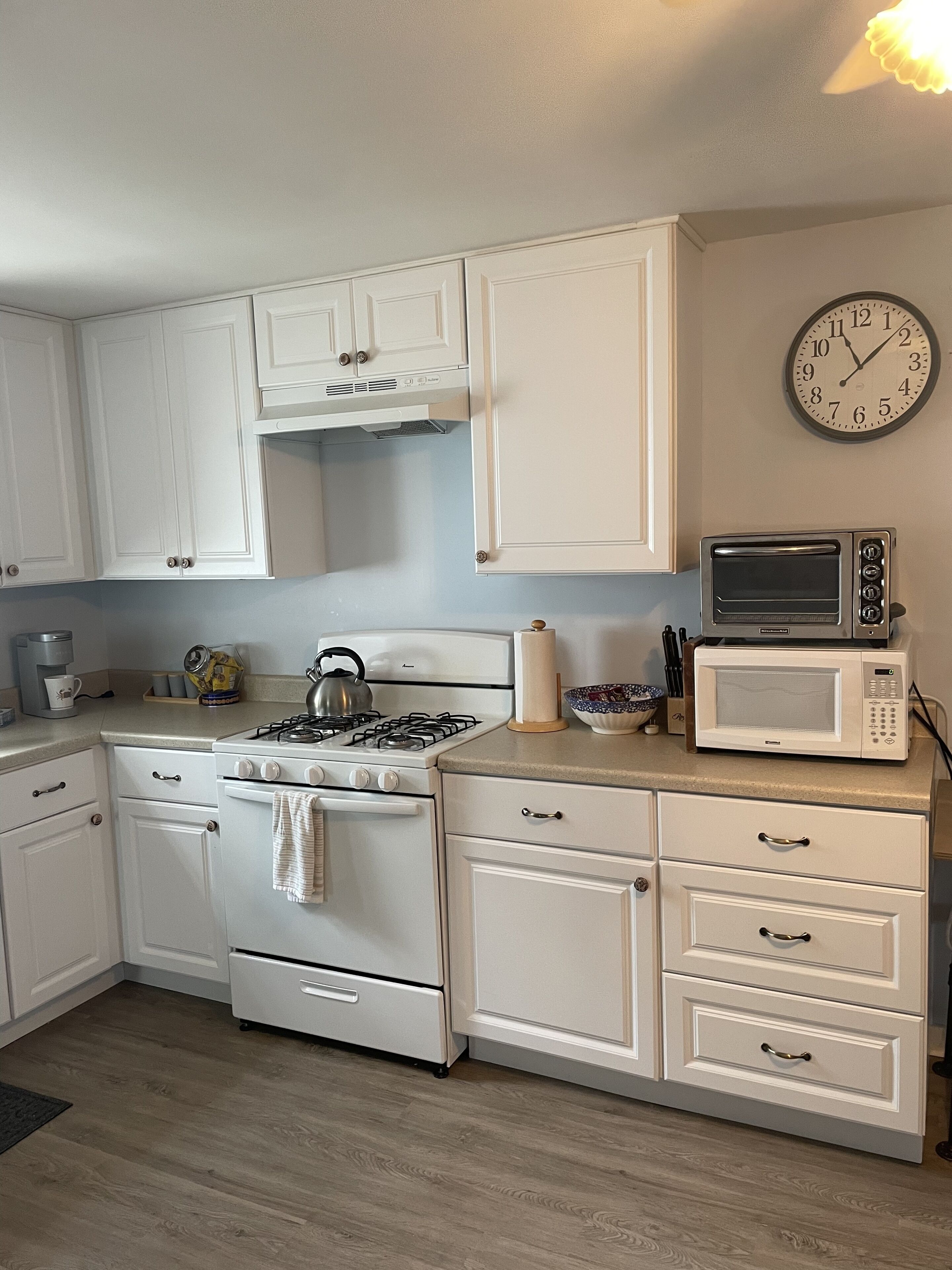 A kitchen with white cabinets, a gas stove, a microwave, and a clock on the wall.