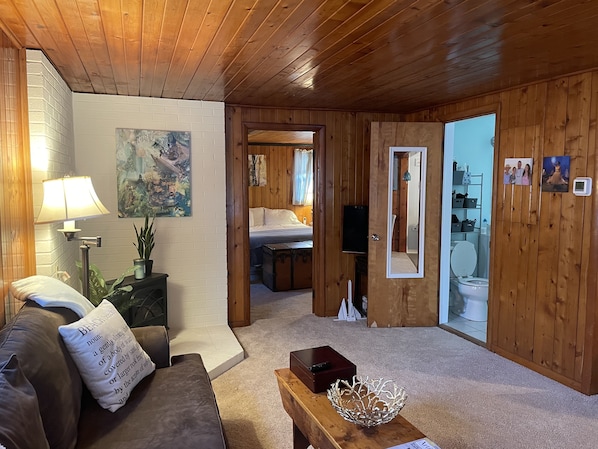 A cozy room with wooden walls, a bed, a sitting area with a couch and a coffee table, and a bathroom visible through an open door.