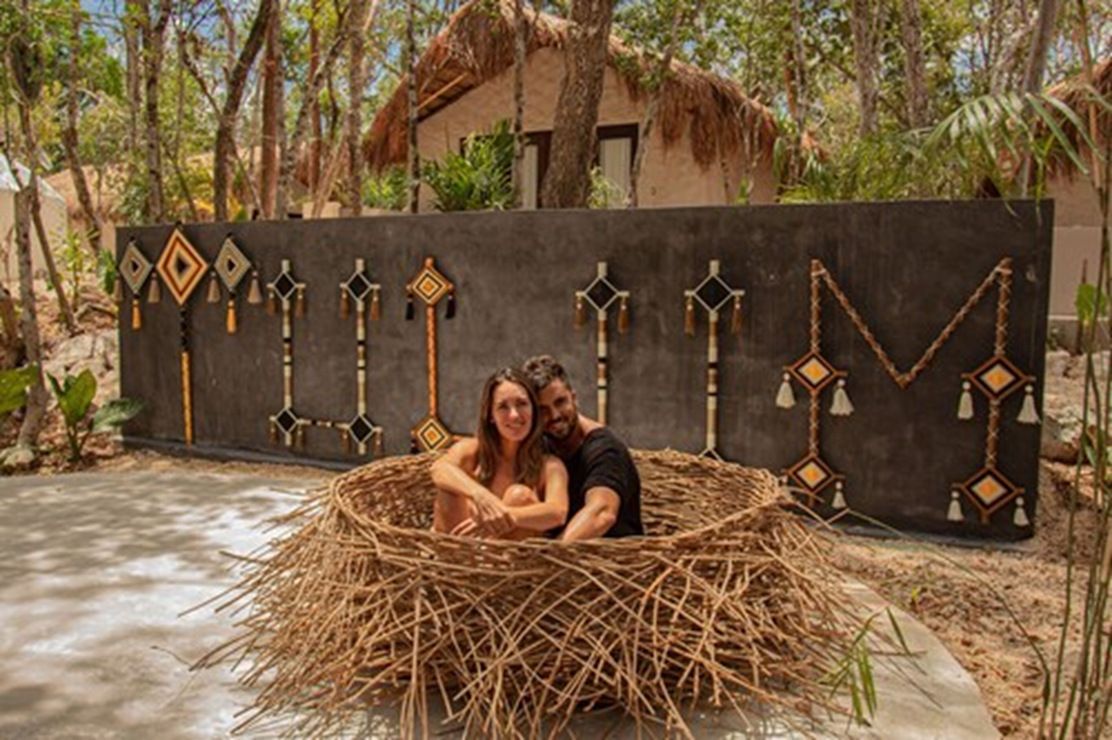A couple sitting in a thatched structure with a decorative wall featuring geometric patterns in the background.