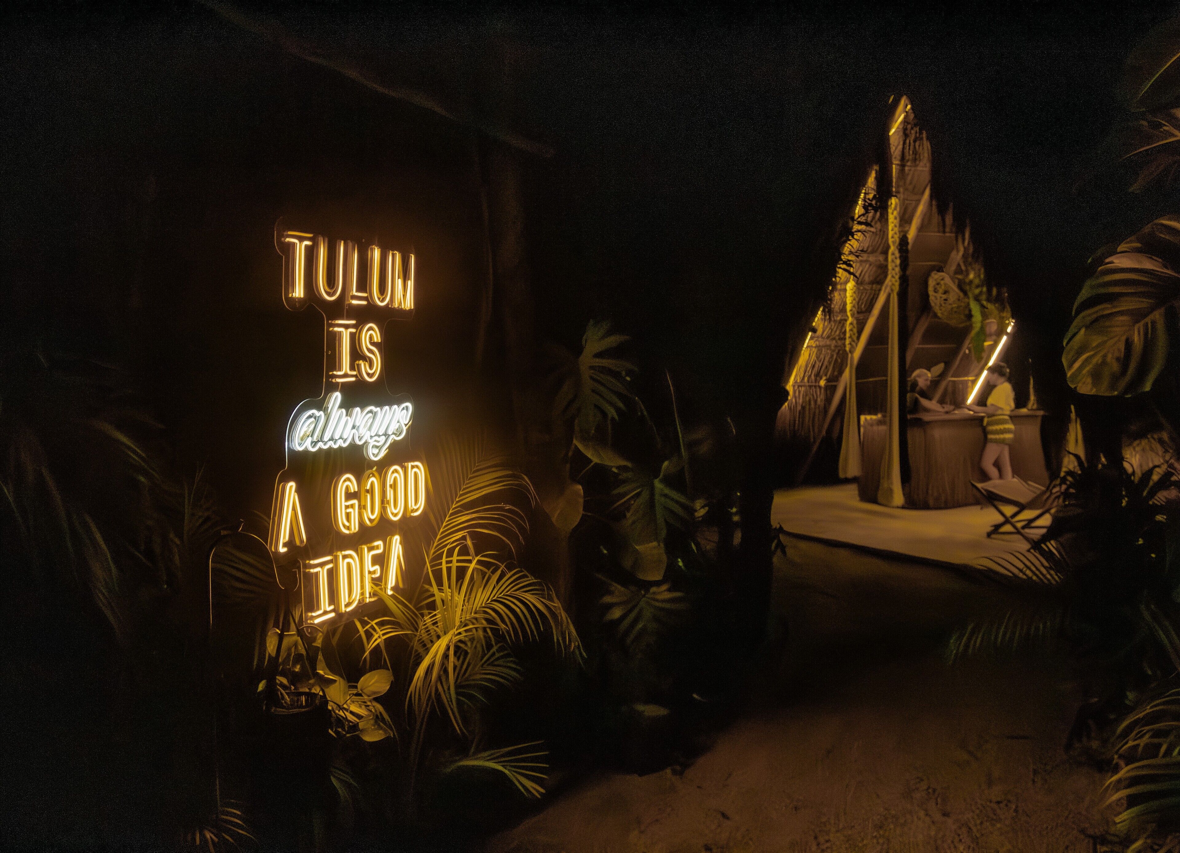 A neon sign with the text "TULUM IS ALWAYS A GOOD IDEA" in a tropical setting with plants and a tiki bar.