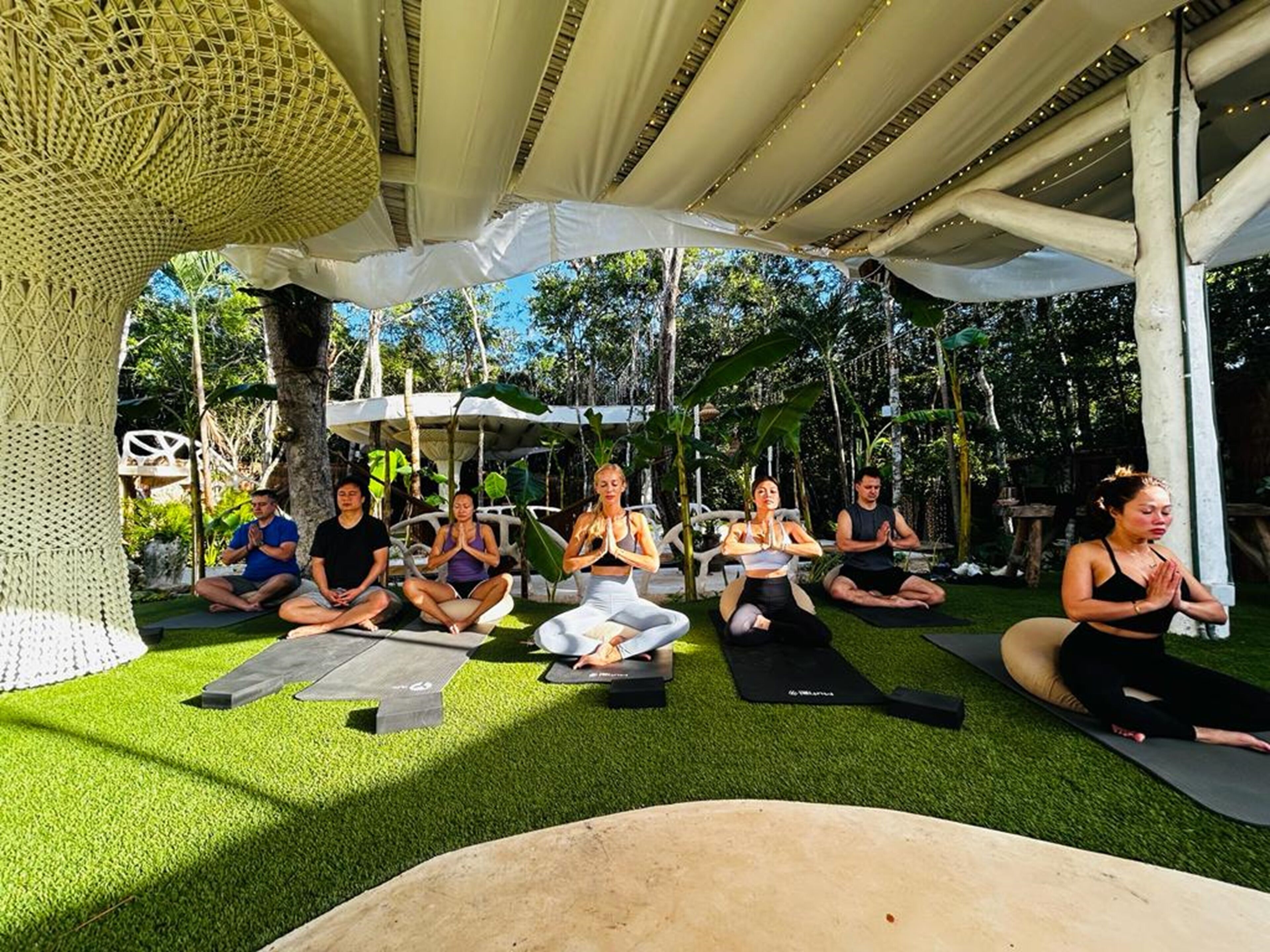 Un gruppo di persone sedute su tappetini da yoga sotto una tettoia in un ambiente esterno, con vegetazione sullo sfondo.