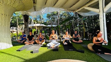 Un gruppo di persone sedute su tappetini da yoga sotto una tettoia in un ambiente esterno, con vegetazione sullo sfondo.