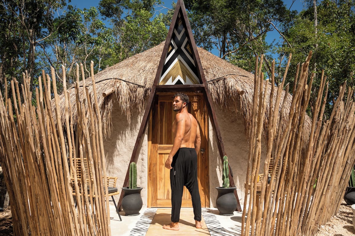 A man standing in front of a thatched-roof hut with a geometric design, surrounded by tall bamboo structures.