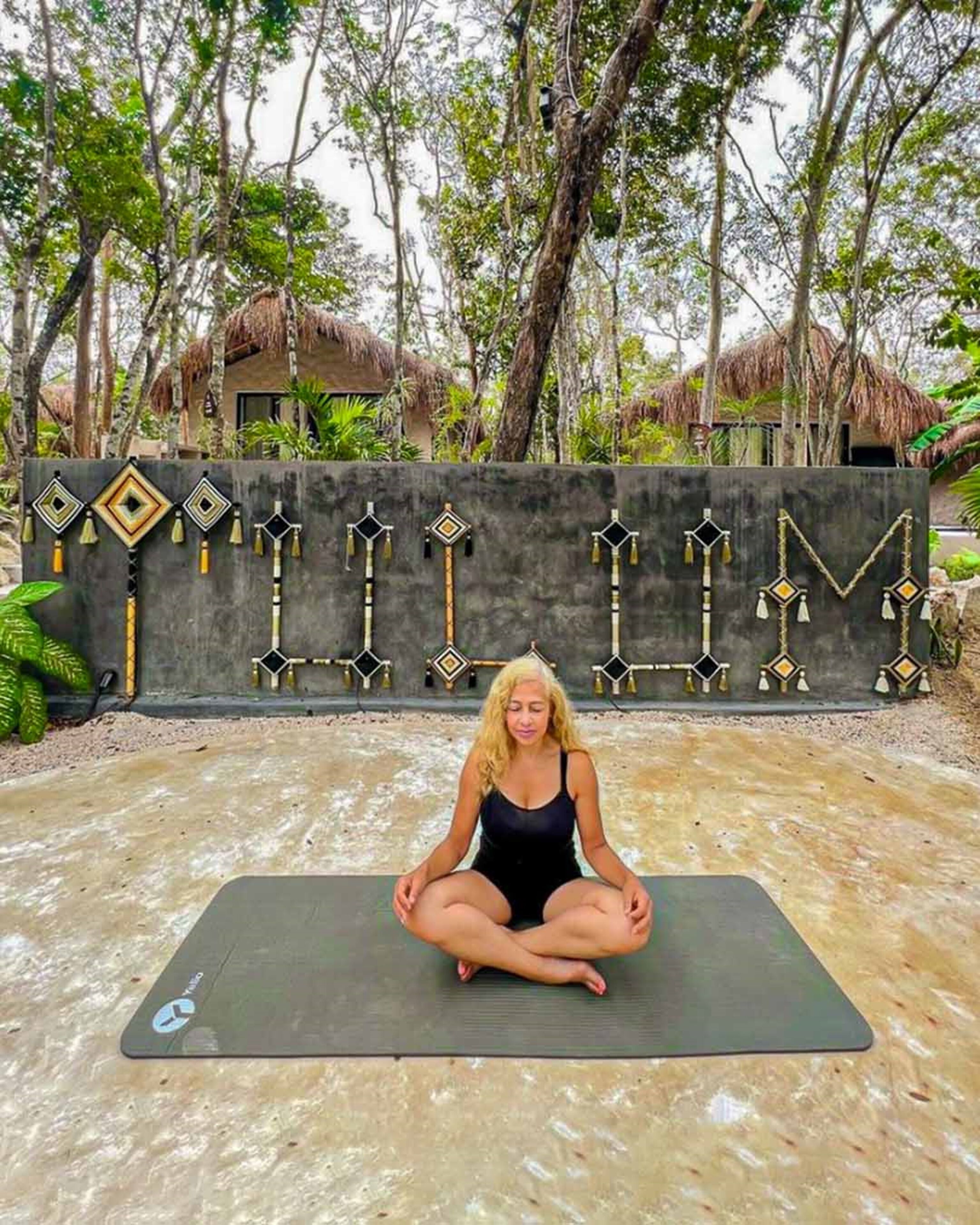 Una persona sentada en una colchoneta de yoga al aire libre, con un muro decorativo al fondo.