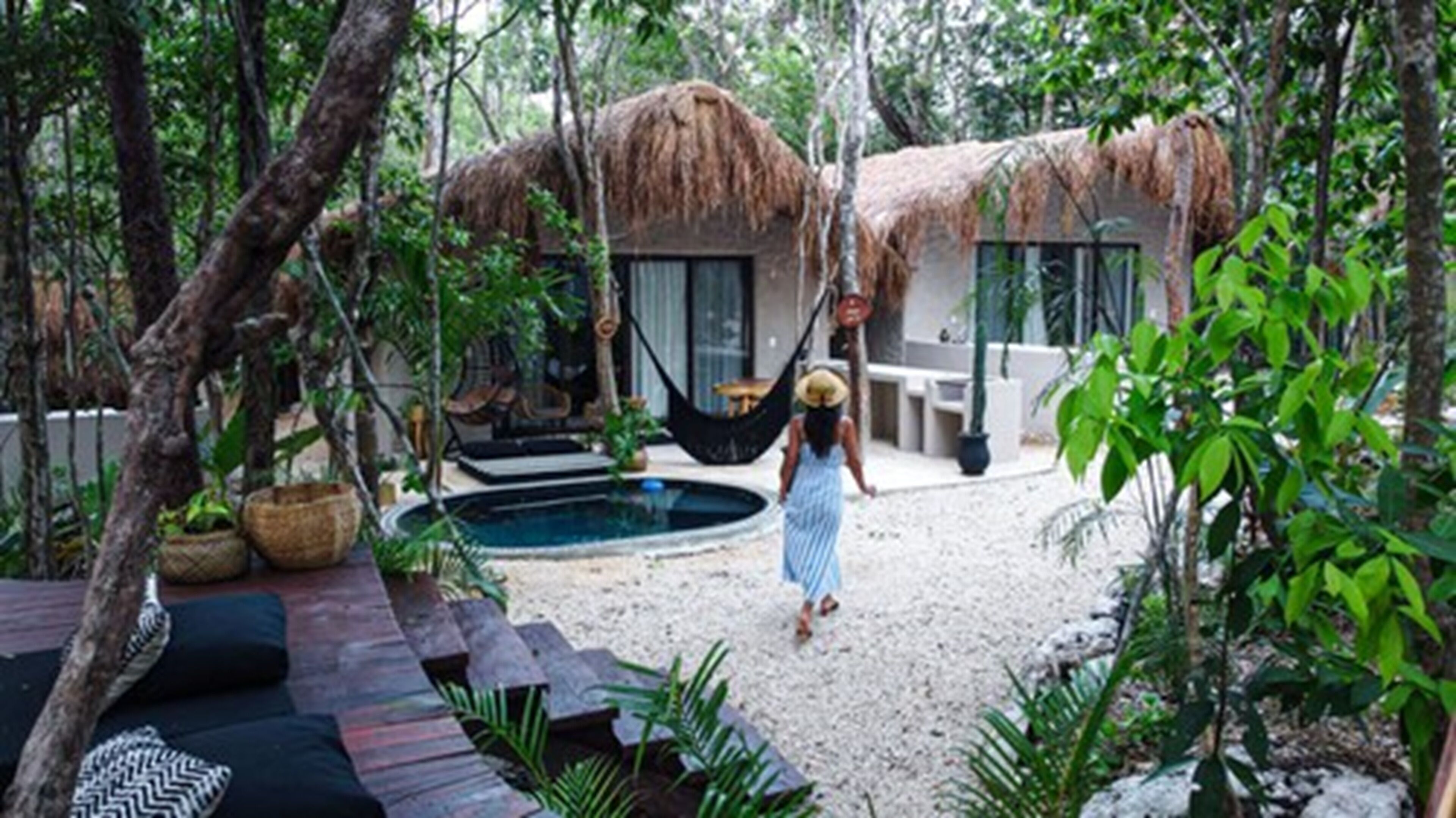 Una casa con techo de paja y piscina, rodeada de vegetación tropical y una hamaca.