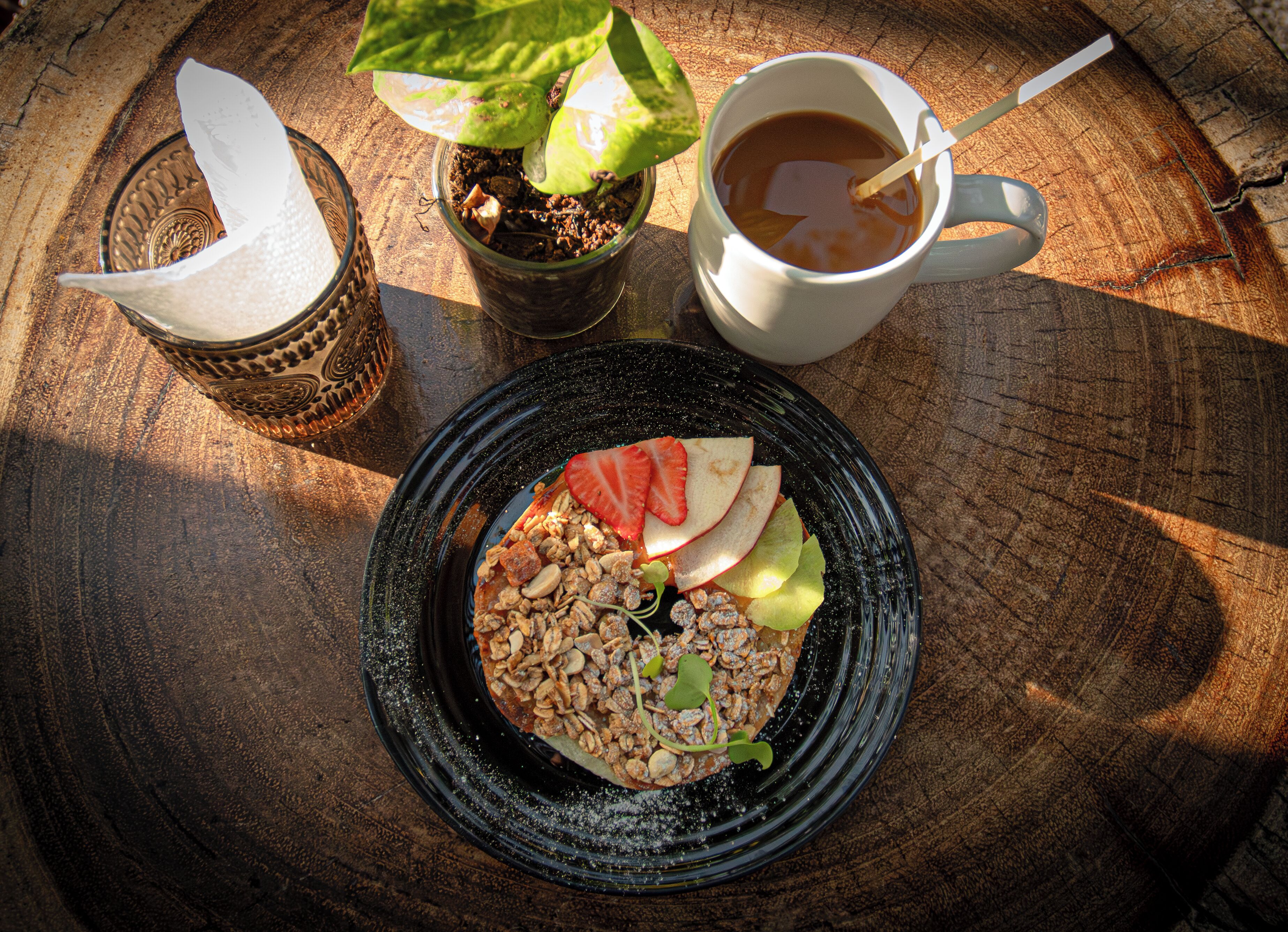 Un desayuno con un plato de granola, frutas y nueces, una taza de café y una pequeña planta en maceta sobre una mesa de madera.