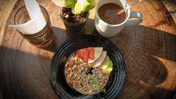 Un desayuno con un plato de granola, frutas y nueces, una taza de café y una pequeña planta en maceta sobre una mesa de madera.