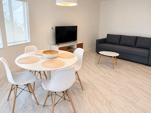 A modern living room with a round dining table, white chairs, a grey sofa, and a flat-screen TV. - The Marlin Beachside (Hollywood)