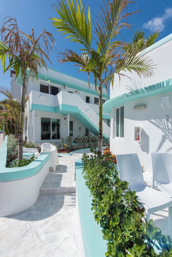 A modern building with a white and turquoise facade, a staircase leading to the upper floor, and outdoor seating areas with white chairs and tables.