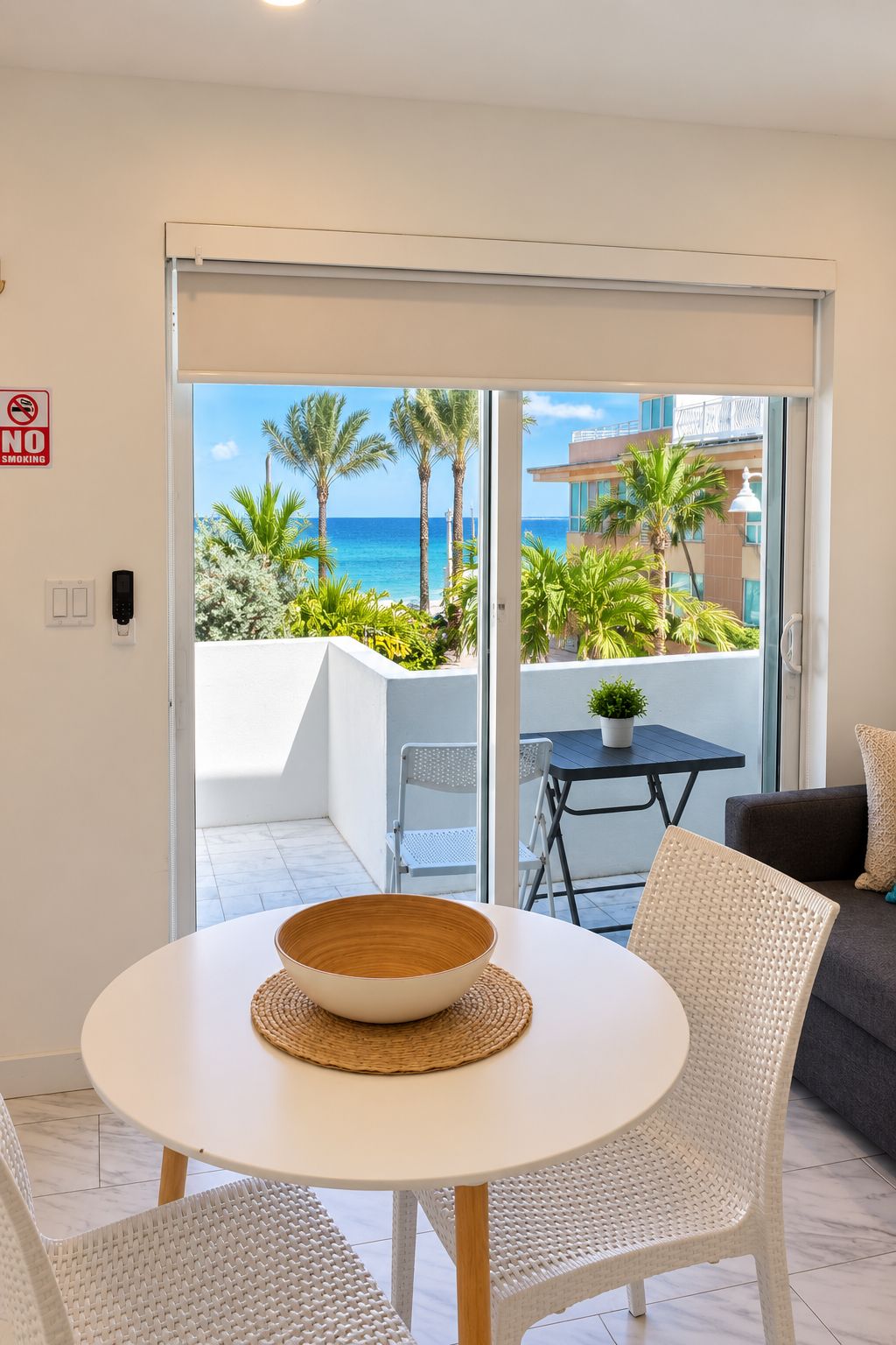 A balcony with a view of the ocean and palm trees, a white table with a bowl, and a wicker chair.