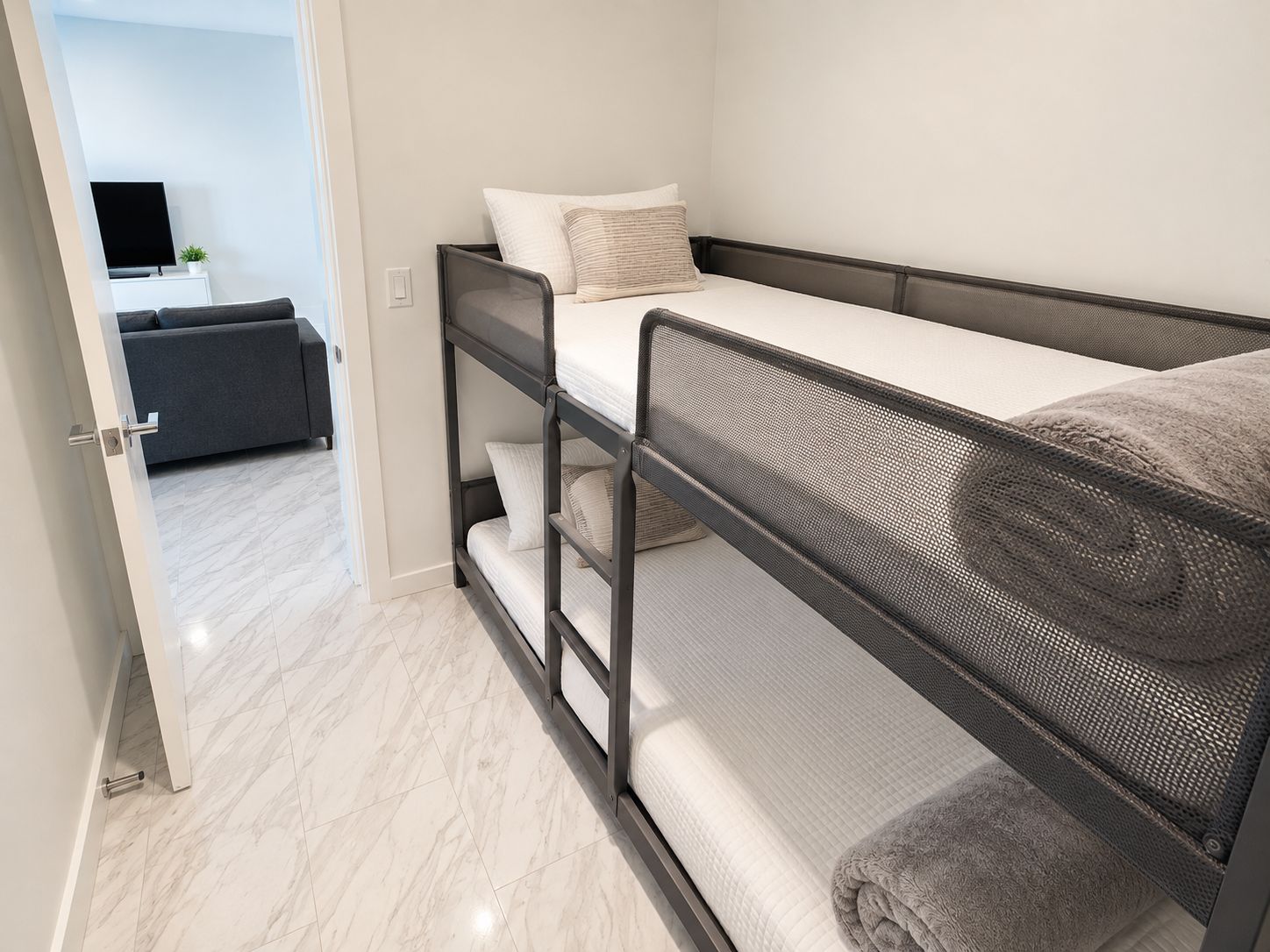 A bunk bed with a mattress and pillows, a television on a stand, and a sofa in a room with white walls and a tiled floor.