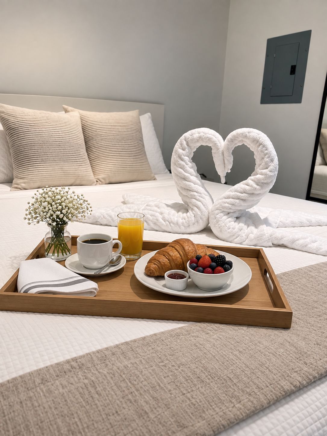 Bandeja de desayuno ordenada con croissants, frutos rojos, café y jugo sobre una cama de hotel con una decoración de toalla con forma de corazón.