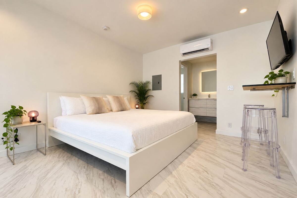 A modern bedroom with a large bed, bedside tables, a TV, and a bathroom visible through an open door.