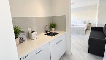 A modern kitchen with a dishwasher, a kettle, and a toaster. There is a sink with a plant beside it. The kitchen leads to a bedroom with a bed and a small table with a plant.
