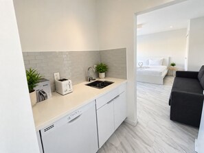 A modern kitchen with a dishwasher, a kettle, and a toaster. There is a sink with a plant beside it. The kitchen leads to a bedroom with a bed and a small table with a plant. - The Marlin Beachside (Hollywood)