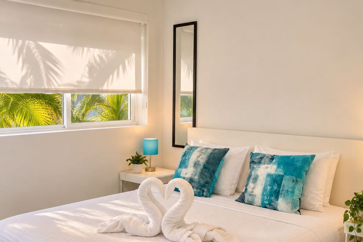 A modern bedroom with a large bed, white bedding, and a tropical view through the window.