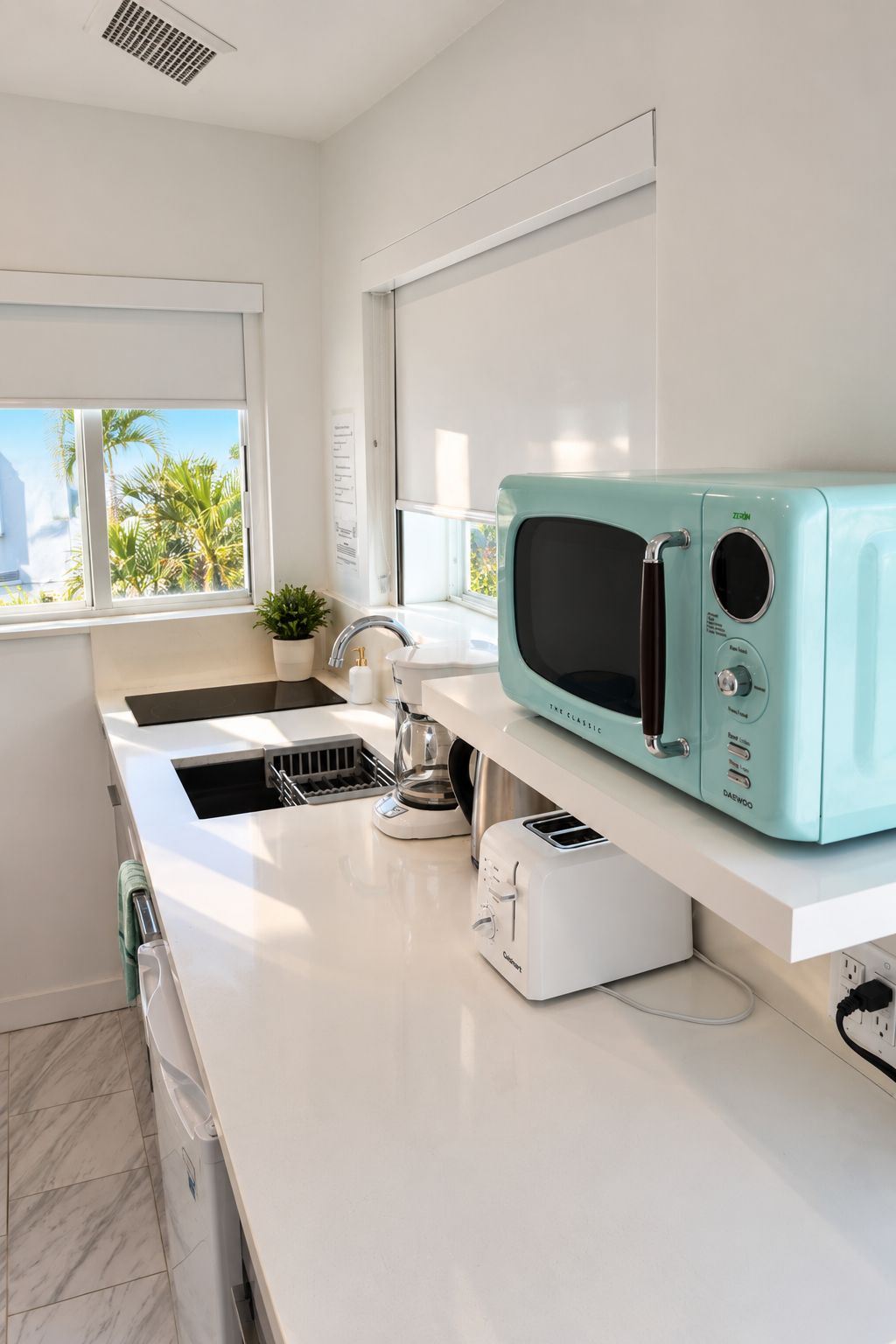 Una cocina moderna con microondas color turquesa, encimeras blancas, un lavamanos y una ventana con vistas a palmeras.