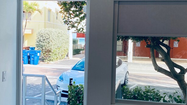 A view through a window of a street with parked cars, a tree, and a building with a red wall. - The Marlin Beachside (Hollywood)