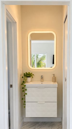 A bathroom with a white vanity, a mirror with a lighted frame, a plant, and a door. - The Marlin Beachside (Hollywood)