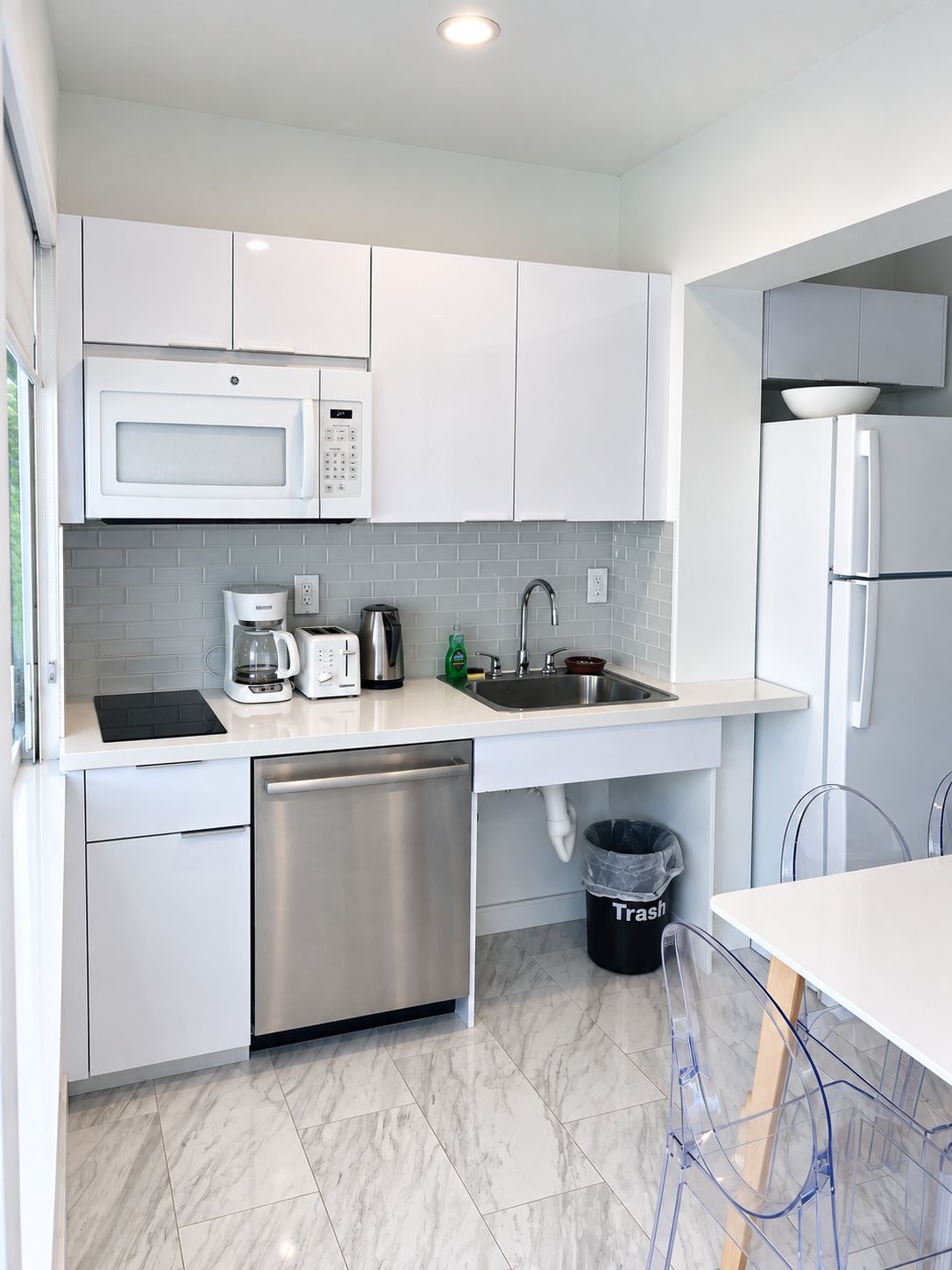 Una cocina compacta con armarios blancos, lavavajillas de acero inoxidable, microondas, cafetera y un cubo de basura.