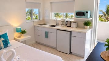 Habitación de hotel compacta con kitchenette, una cama con almohada azul, lámpara de noche y vistas a palmeras.
