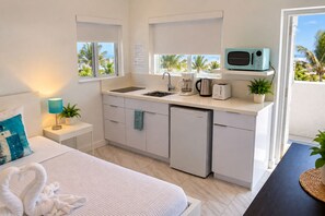 A compact hotel room with a kitchenette, a bed with a blue pillow, a bedside lamp, and a view of palm trees. - The Marlin Beachside (Hollywood)
