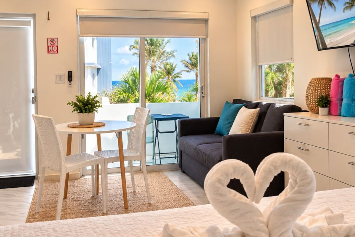 A hotel room with a bed, a sofa, a dining area with chairs, a TV, and a view of the beach.