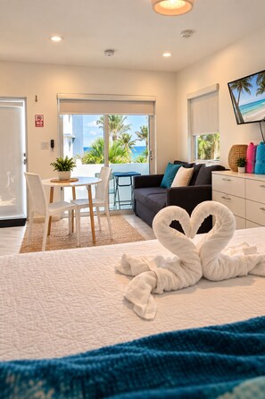 A hotel room with a bed, a sofa, a dining area with chairs, a TV, and a view of the beach. - The Marlin Beachside (Hollywood)