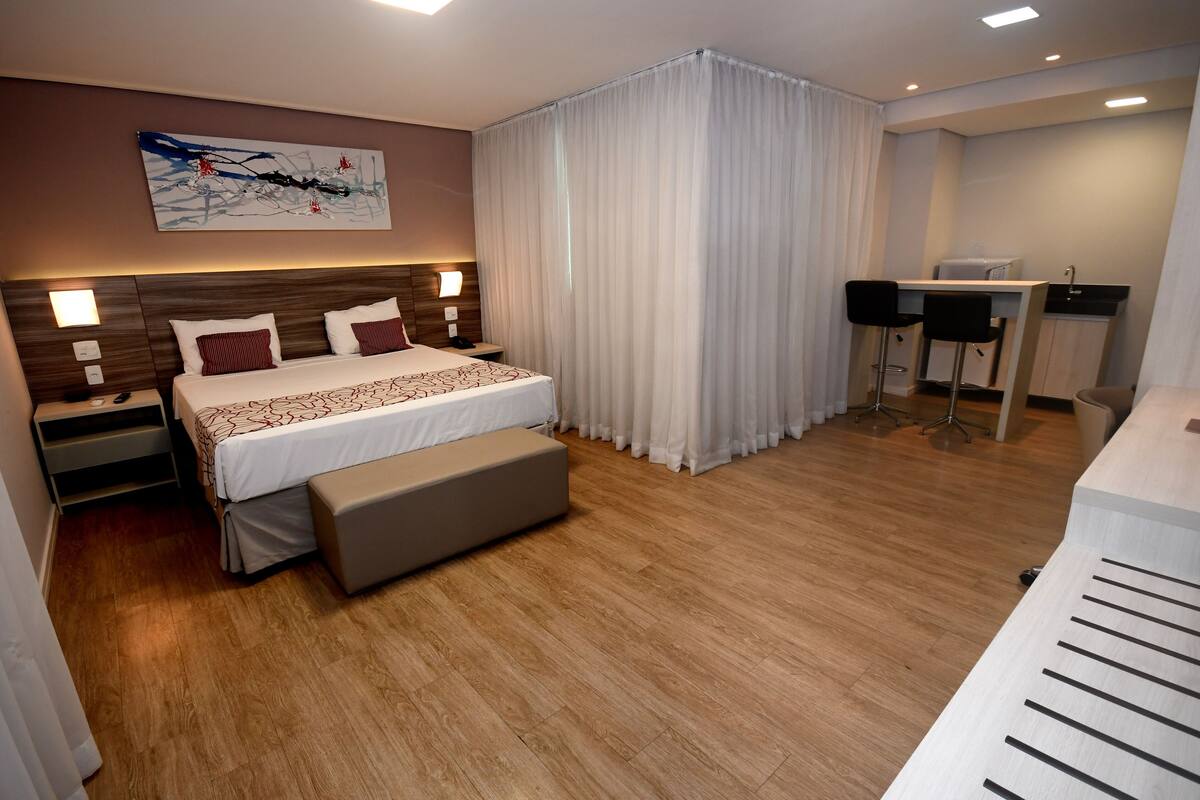 Una habitación de hotel moderna con una cama grande, mesitas de noche, un escritorio con dos sillas y una kitchenette pequeña.